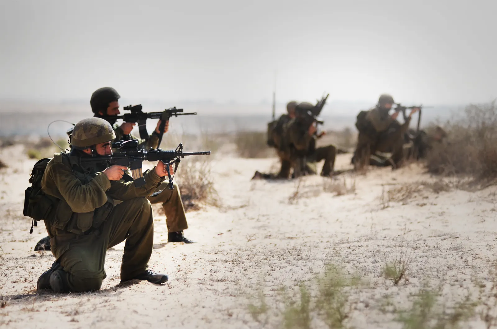 Cinco soldados israelíes arrodillados sobre terreno arenoso, con casco y fusil de asalto apuntando hacia el horizonte durante un ejercicio militar