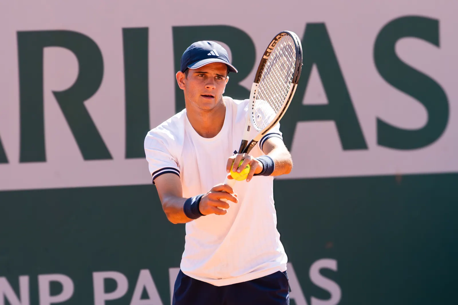 Rafael Jódar, con camiseta blanca, gorra azul marino y muñequeras, sujeta la pelota contra el cordaje de la raqueta antes de sacar, delante de una lona publicitaria de BNP Paribas.