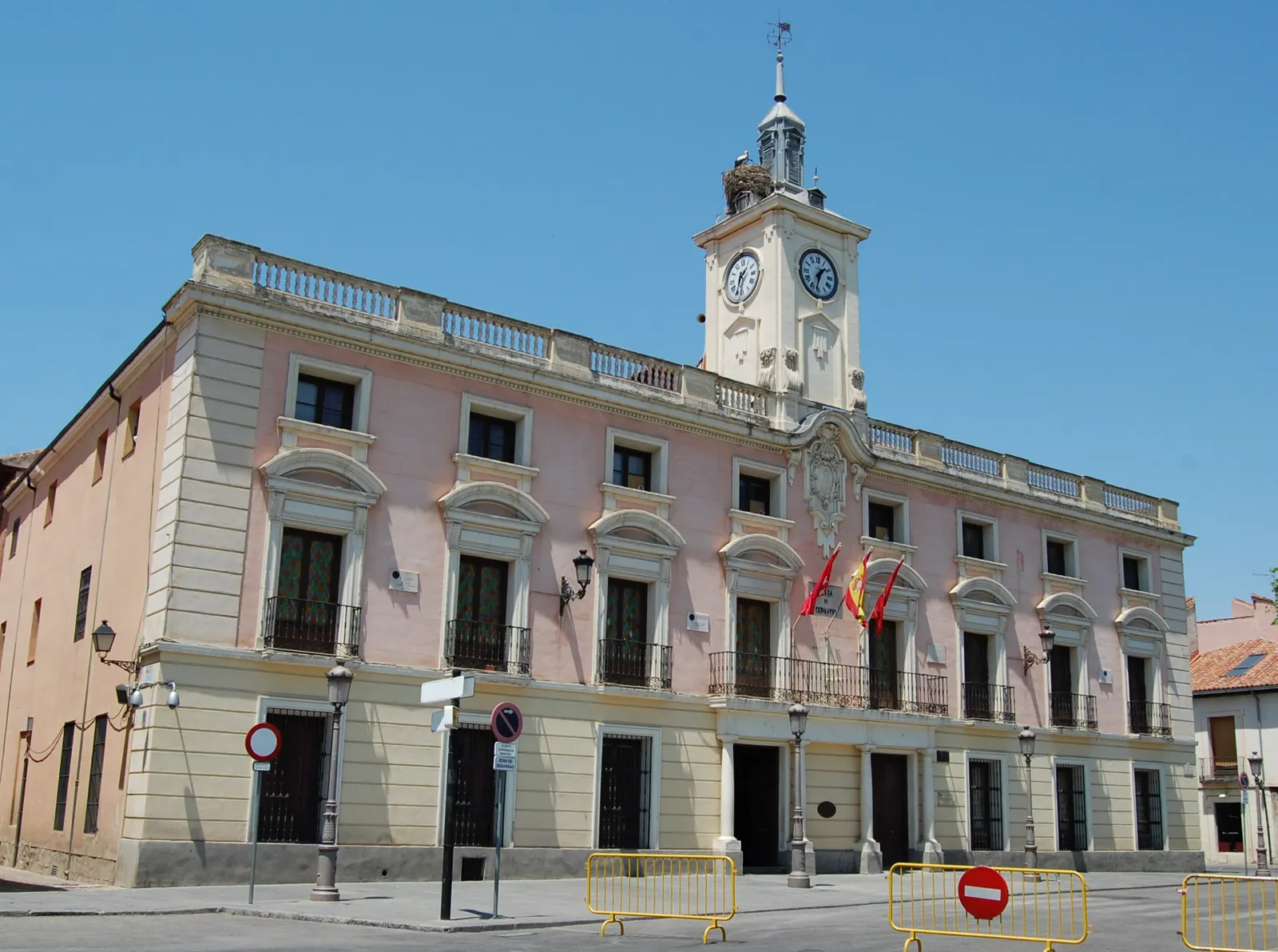 Fachada principal del Ayuntamiento de Alcalá de Henares, edificio de piedra y ladrillo con arcos en la planta baja, balcones corridos y una torre con reloj rematada por un campanil, bajo cielo despejado.