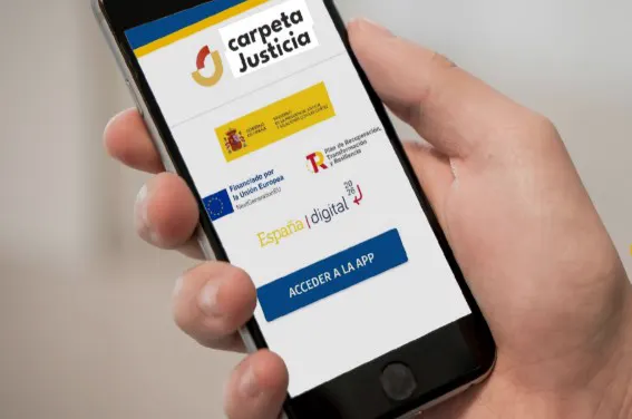 Una mano sostiene un teléfono móvil con la pantalla de acceso a la aplicación Carpeta Justicia, con su logotipo, los escudos del Ministerio de la Presidencia, Justicia y Relaciones con las Cortes, los emblemas de los fondos europeos y un botón azul que dice «Acceder a la app».