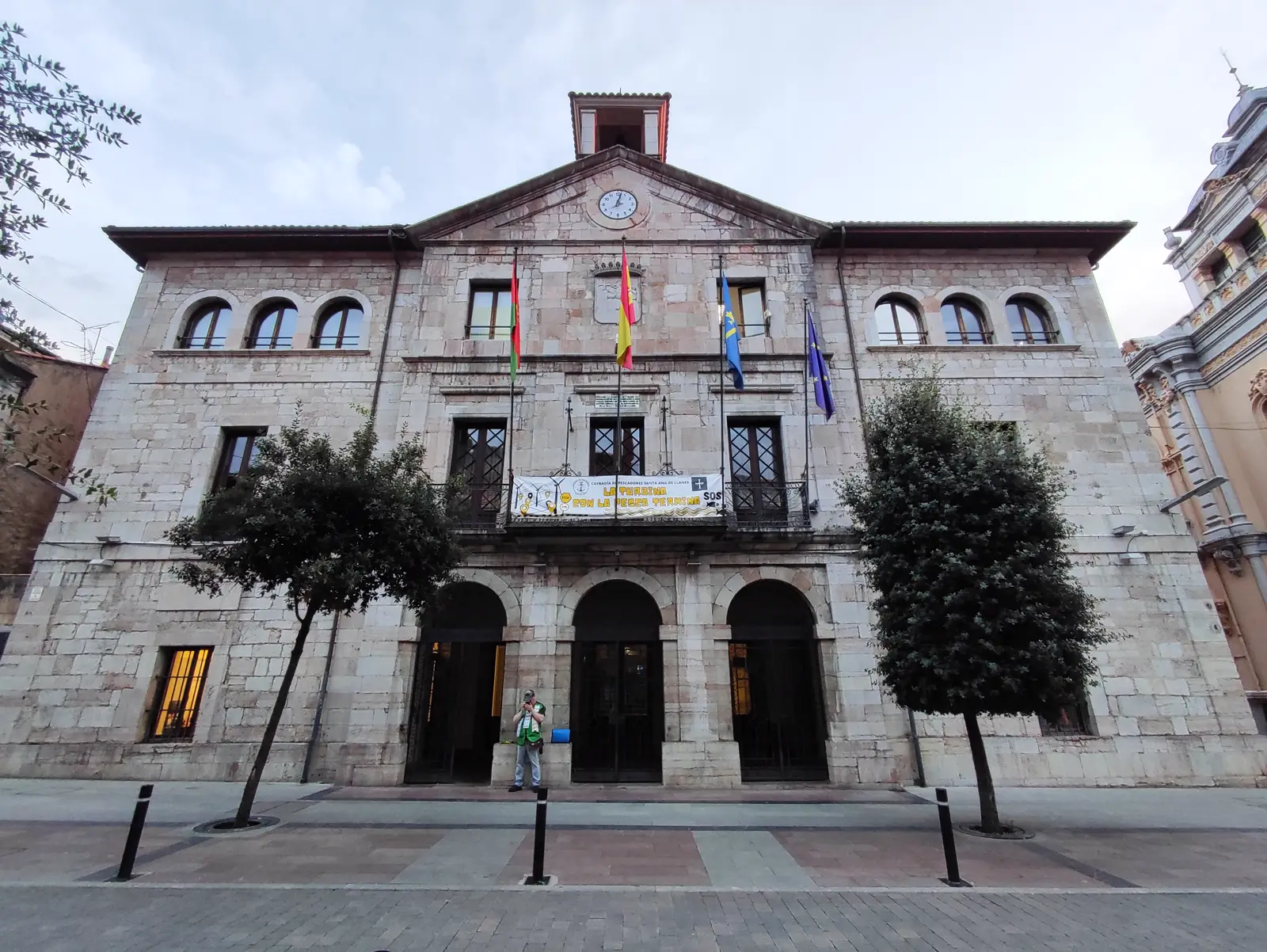 Fachada de piedra de un edificio consistorial de tres alturas con soportal de tres arcos, reloj en el frontón y cuatro banderas en el balcón central, con dos árboles delante