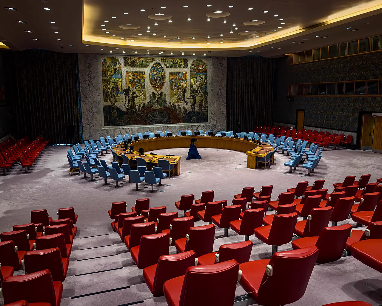 Sala vacía del Consejo de Seguridad de la ONU, con la mesa en herradura, las butacas azules de las delegaciones, las rojas del público y el mural de Per Krohg al fondo.