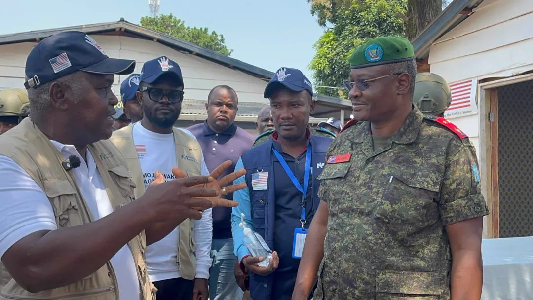 Un hombre con chaleco caqui gesticula mientras habla con un oficial del ejército congoleño de boina verde, rodeados de sanitarios y responsables con chalecos y gorras de la respuesta al ébola