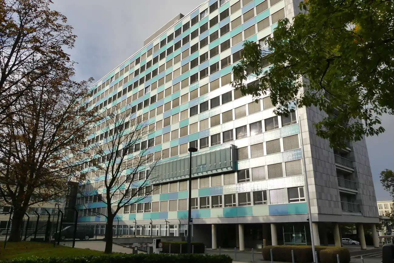 Fachada acristalada de un edificio administrativo de trece plantas con paneles en tonos verdes y grises, rodeado de árboles, bajo un cielo nublado.
