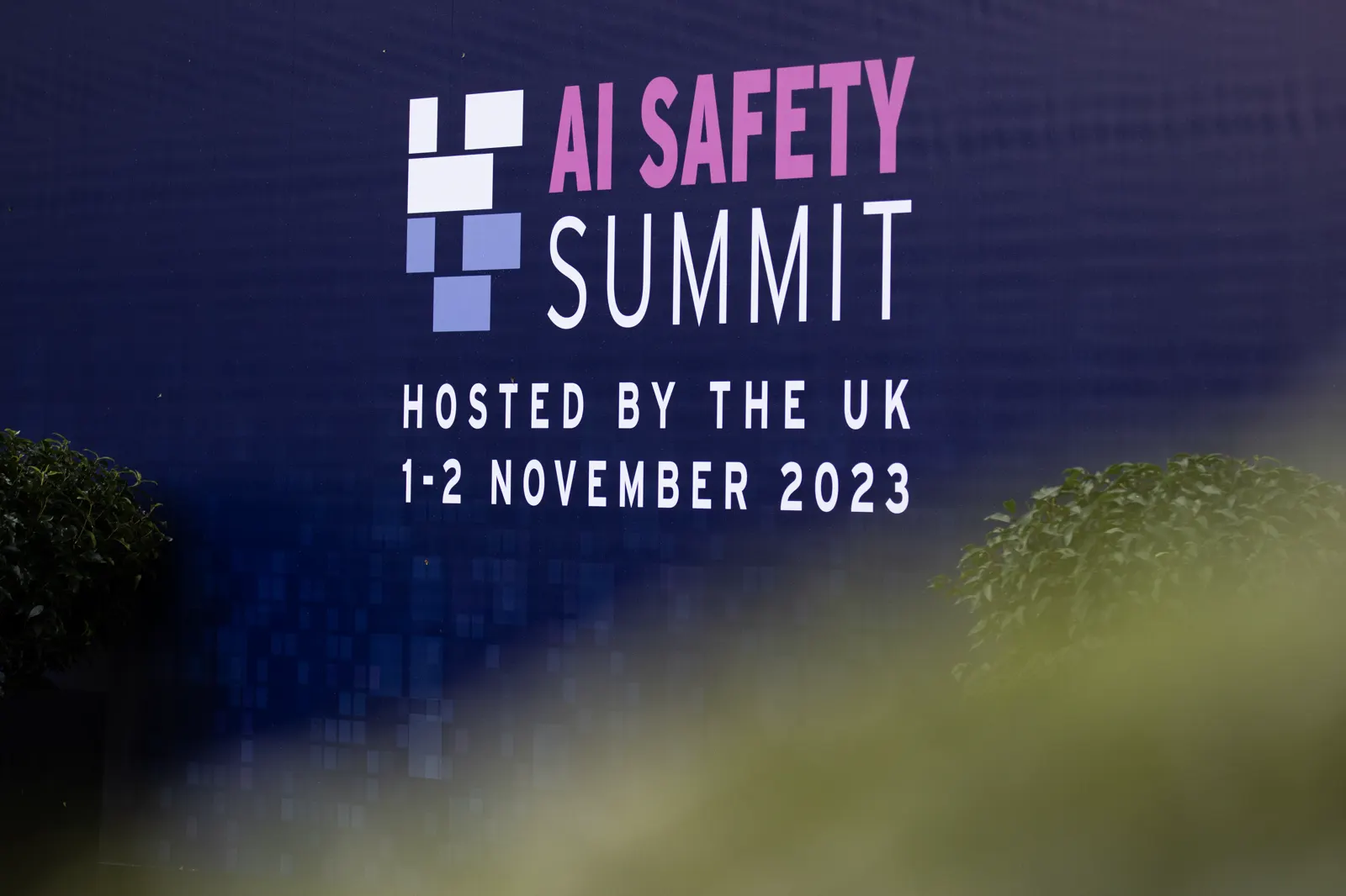 Valla publicitaria azul con el logotipo de la AI Safety Summit celebrada en el Reino Unido el 1 y 2 de noviembre de 2023, rodeada de setos verdes.