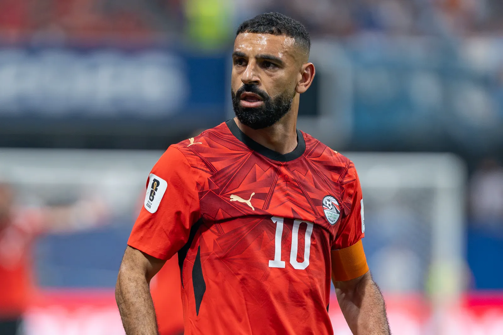 Mohamed Salah, con la camiseta roja de la selección de Egipto, el dorsal 10 y el brazalete naranja de capitán, mira hacia su izquierda sobre el césped con la grada desenfocada al fondo.