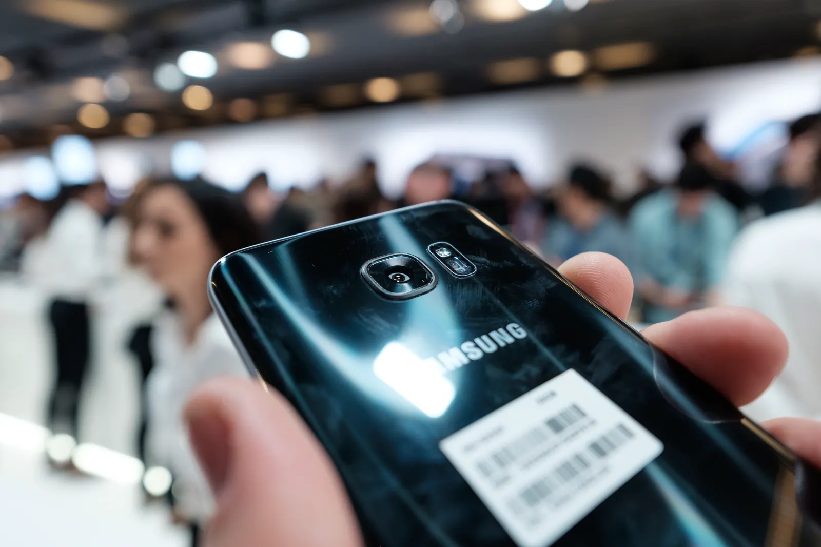 Primer plano de una mano sosteniendo un teléfono móvil negro con el logotipo Samsung en la carcasa trasera, con asistentes desenfocados al fondo en una sala de presentación.