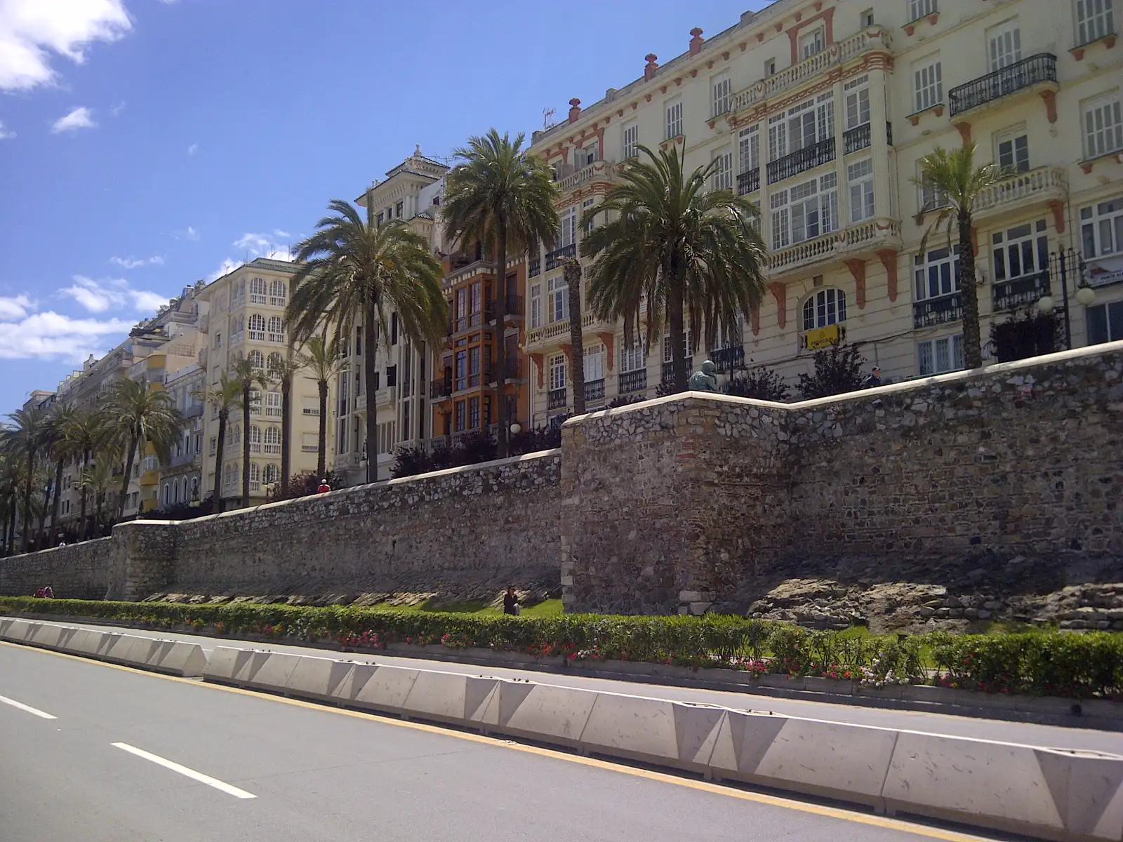 Avenida vacía en el centro de Ceuta junto a un lienzo de muralla de piedra con un seto de flores a sus pies; detrás, una hilera de palmeras y edificios señoriales de fachadas claras con miradores acristalados y balcones de hierro, bajo un cielo azul con nubes.