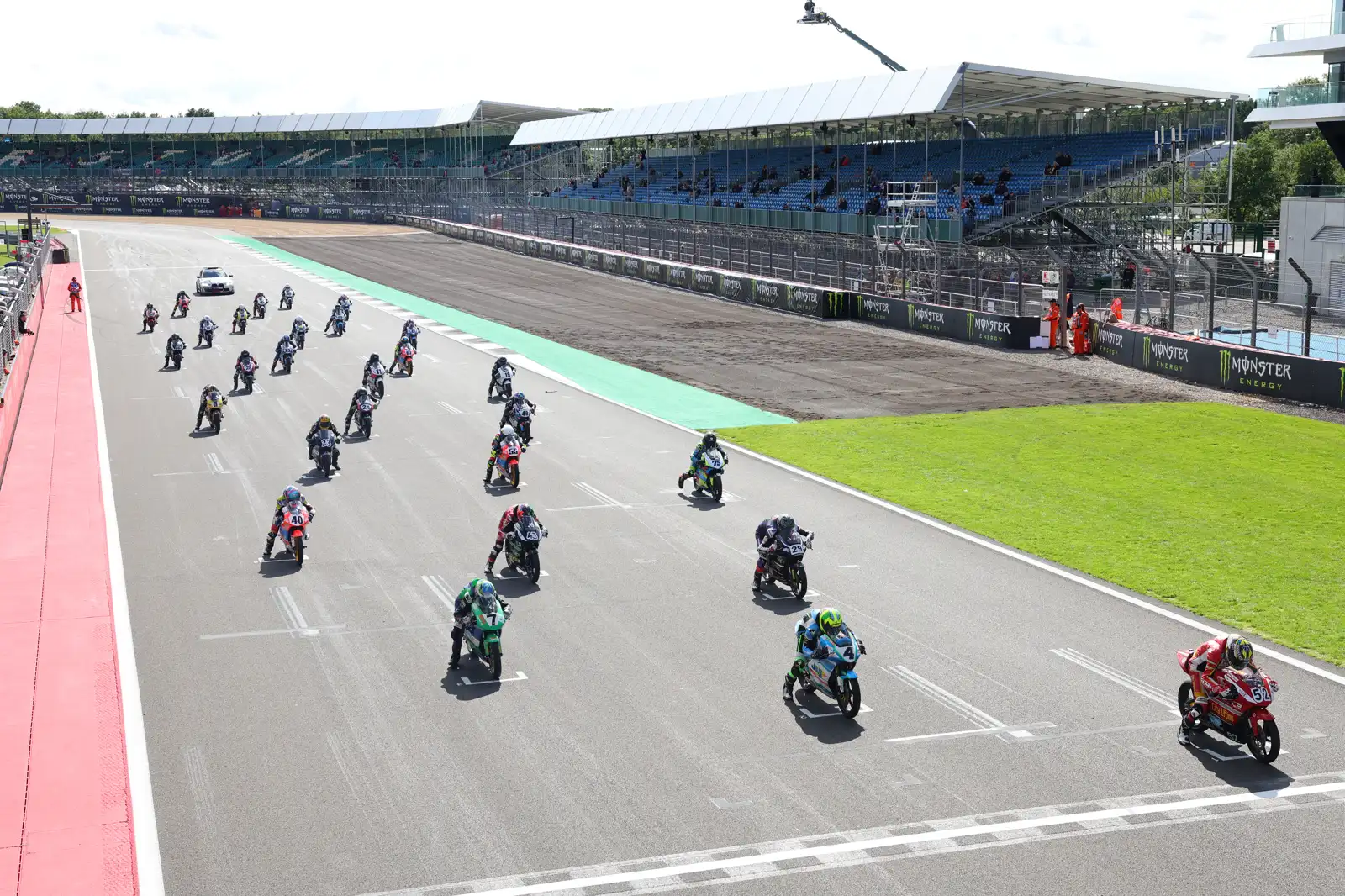 Una parrilla de motos de competición arranca por la recta principal del circuito de Silverstone, con las gradas y los boxes al fondo.