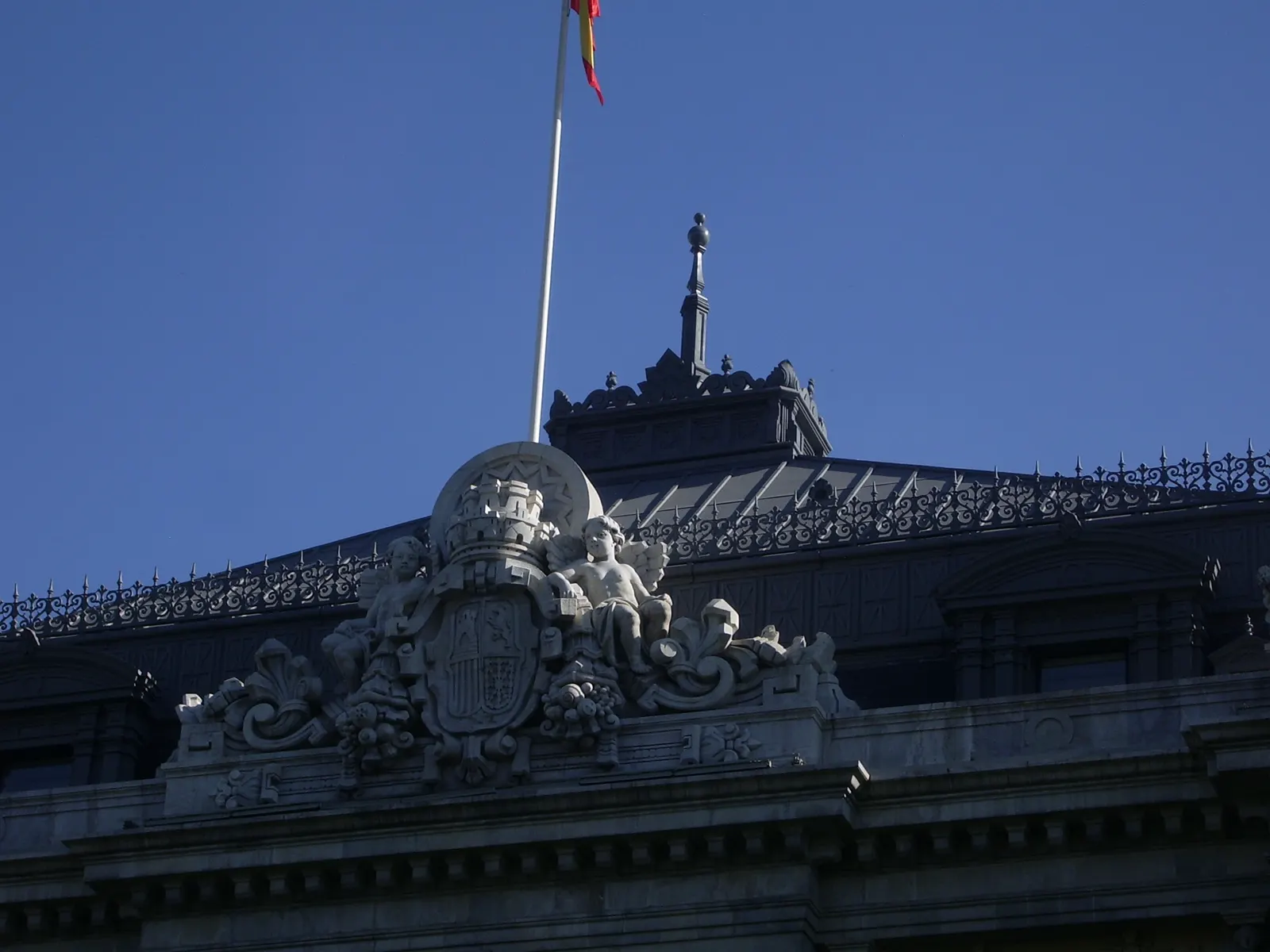 Remate de la cornisa del edificio del Banco de España en Madrid: un grupo escultórico en piedra con el escudo de España y una figura infantil, tras una balaustrada de hierro, con una bandera española ondeando sobre un cielo azul intenso.