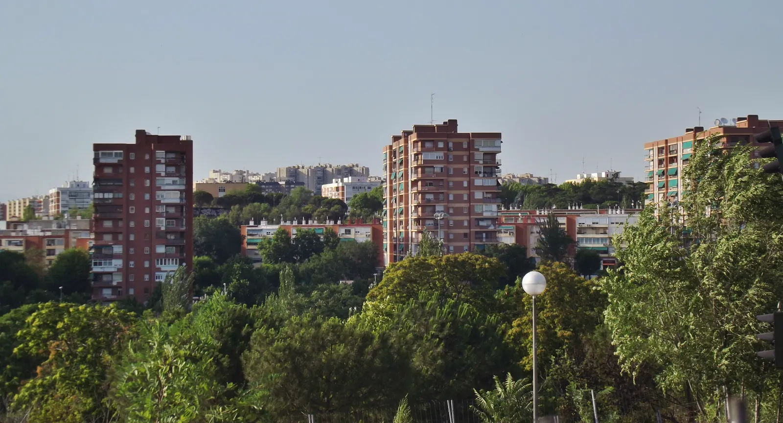Varios bloques altos de viviendas de ladrillo rojo, con balcones y toldos, asoman entre arbolado urbano bajo un cielo despejado en un barrio de Madrid.