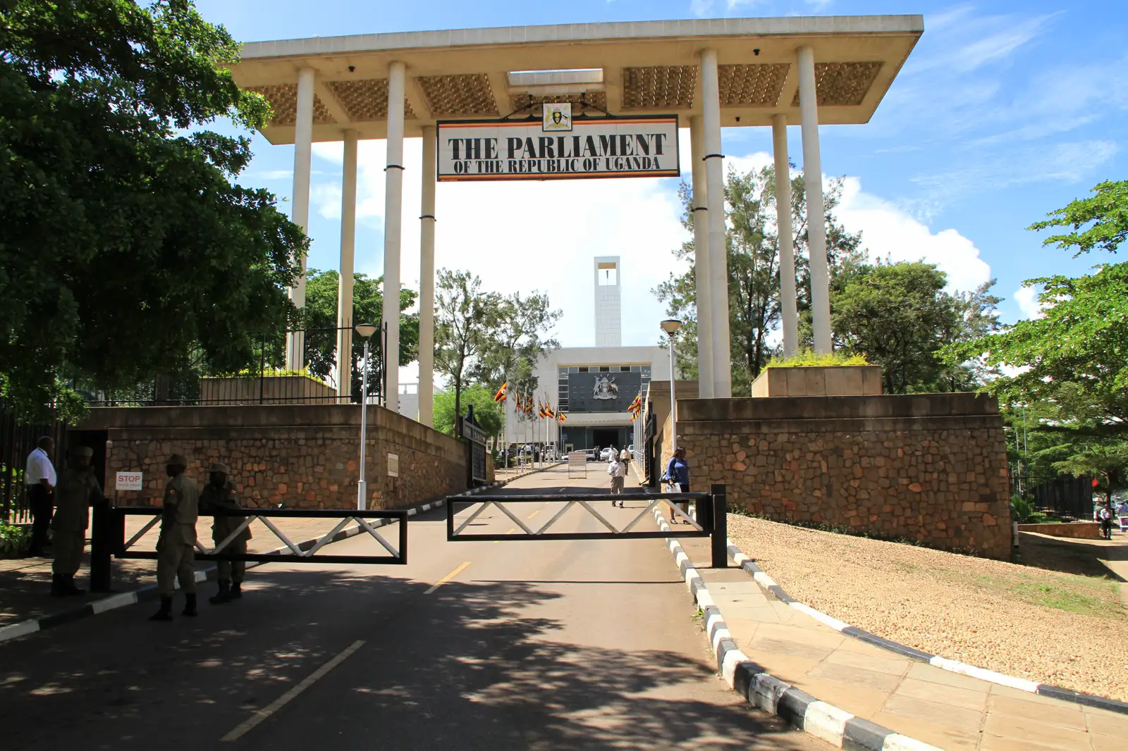 Entrada del recinto del Parlamento de Uganda en Kampala: un pórtico de hormigón con el rótulo «The Parliament of the Republic of Uganda», guardias uniformados junto a una barrera y banderas ugandesas ante el edificio.