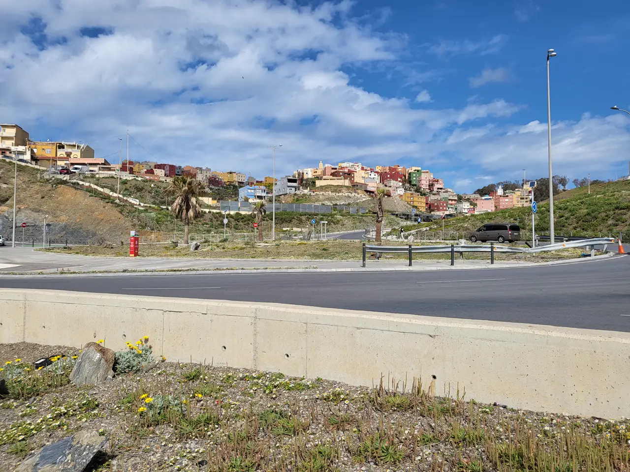 Vista de la barriada de El Príncipe, en Ceuta, desde una carretera: en primer término un muro de hormigón y una rotonda con palmeras; sobre la ladera, casas de colores —amarillas, rojas, rosas y verdes— apiñadas en lo alto del cerro bajo un cielo azul con nubes.