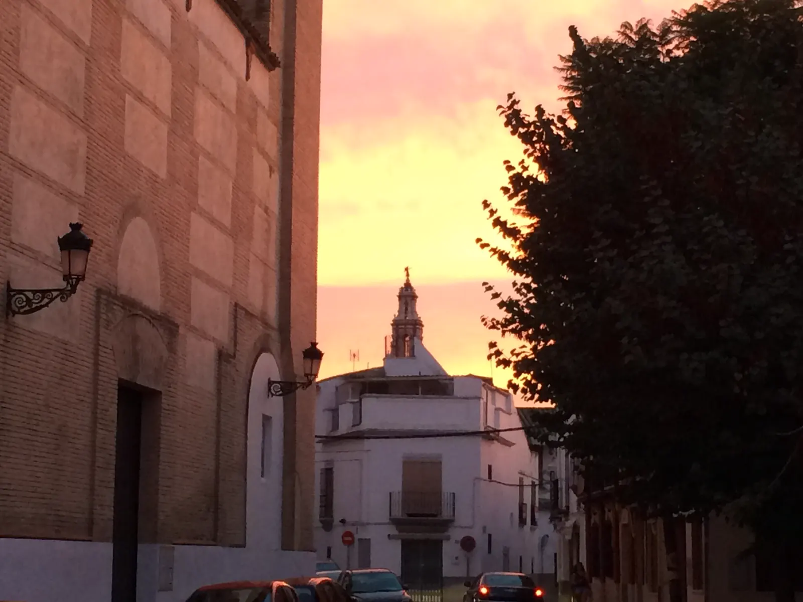 Calle estrecha de Écija con un muro de ladrillo y faroles de forja a la izquierda, casas encaladas al fondo y la torre de una iglesia recortada contra un cielo anaranjado al atardecer.