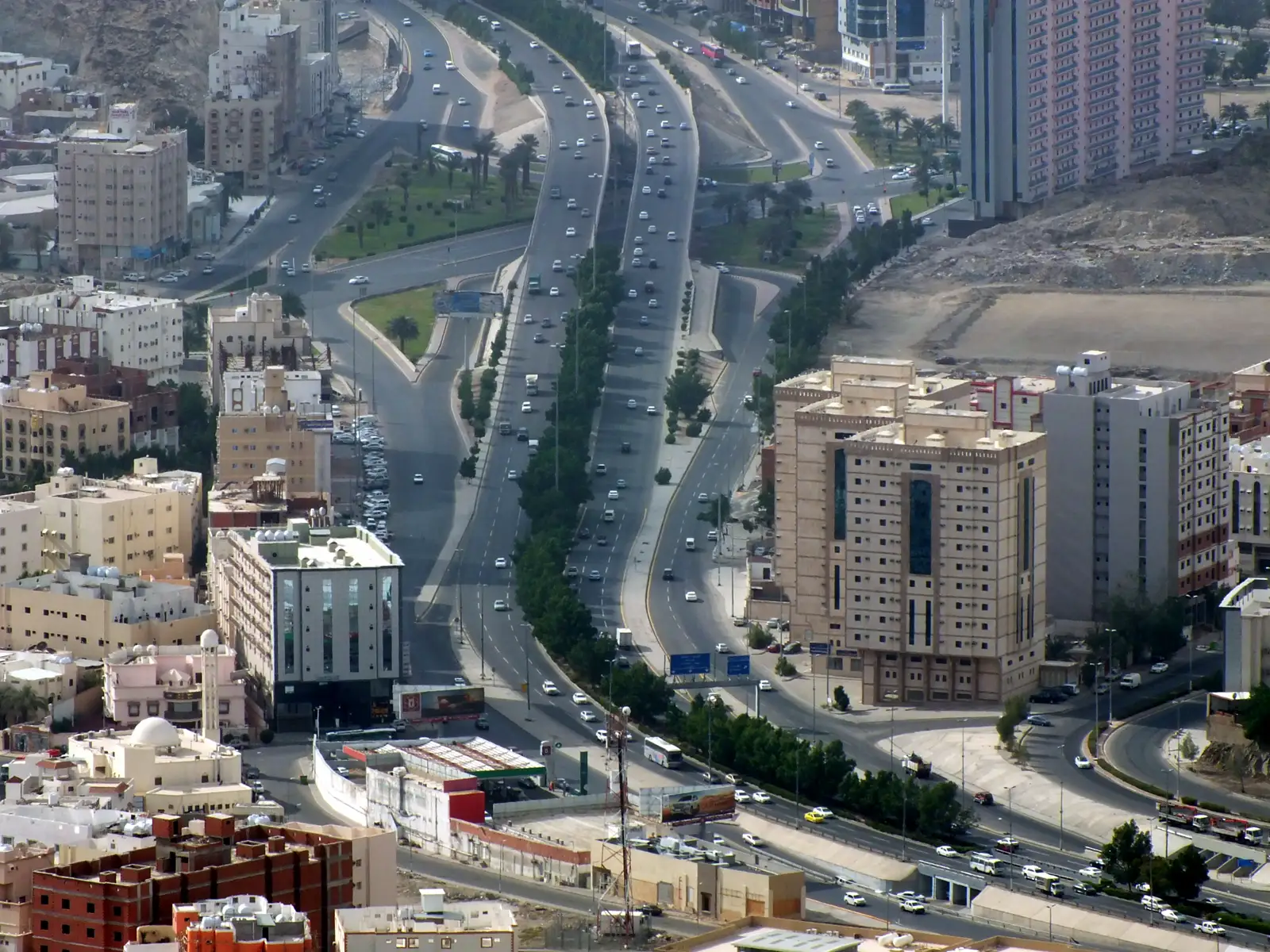 Vista elevada de una avenida de varios carriles con tráfico en La Meca, flanqueada por bloques de viviendas y colinas áridas.