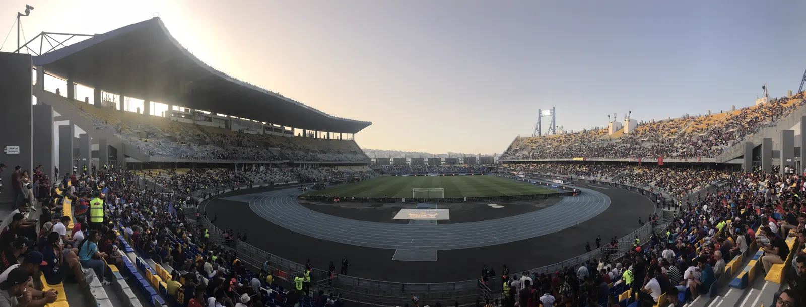 Panorámica del estadio Ibn Battuta de Tánger con las gradas llenas de público, la pista de atletismo azul rodeando el césped y el sol bajo sobre la tribuna principal.