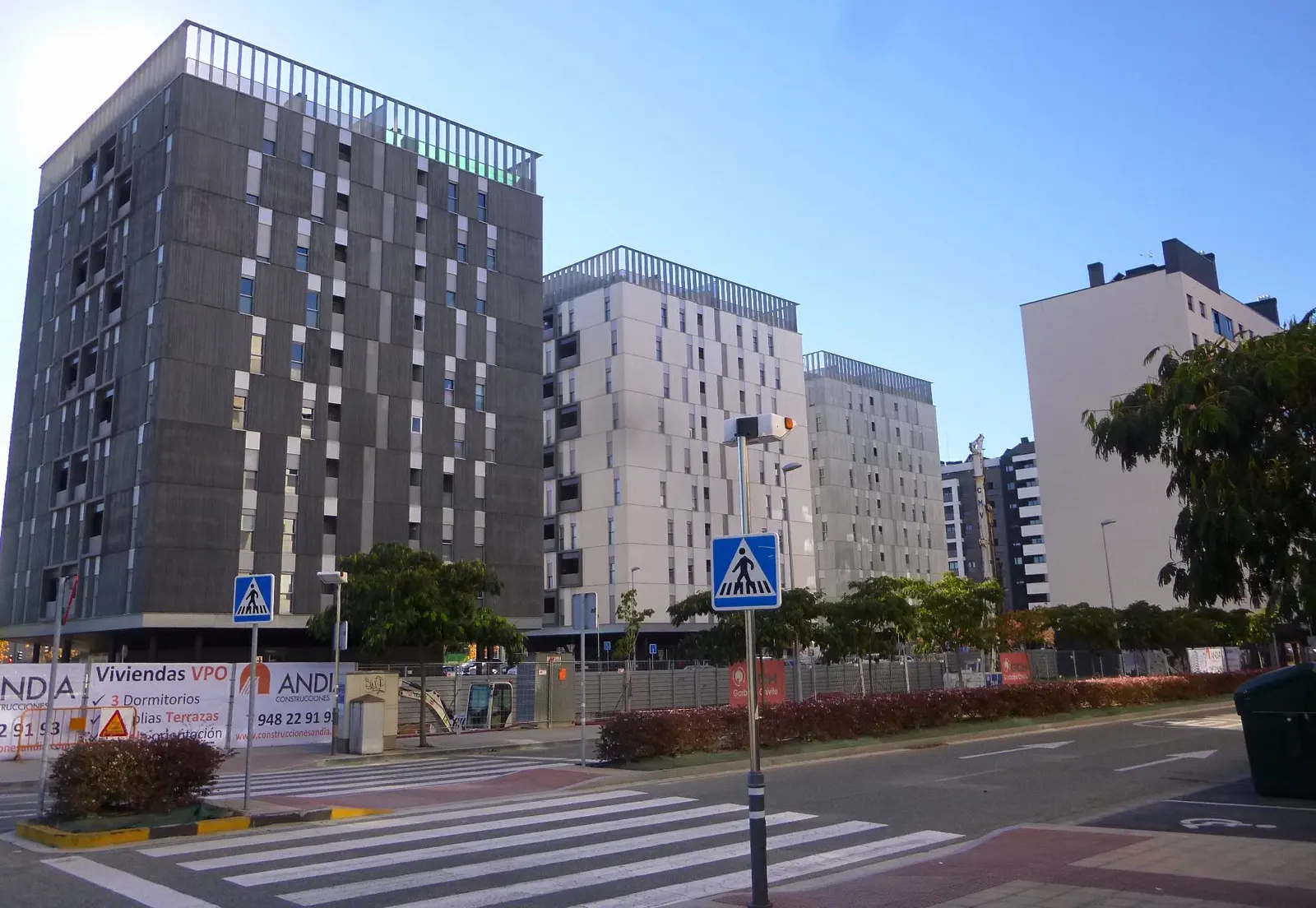 Tres bloques de viviendas de obra reciente, de unas diez alturas y fachadas grises y blancas, junto a una avenida con paso de cebra; en el vallado del solar delantero, un cartel de venta de viviendas de protección oficial.
