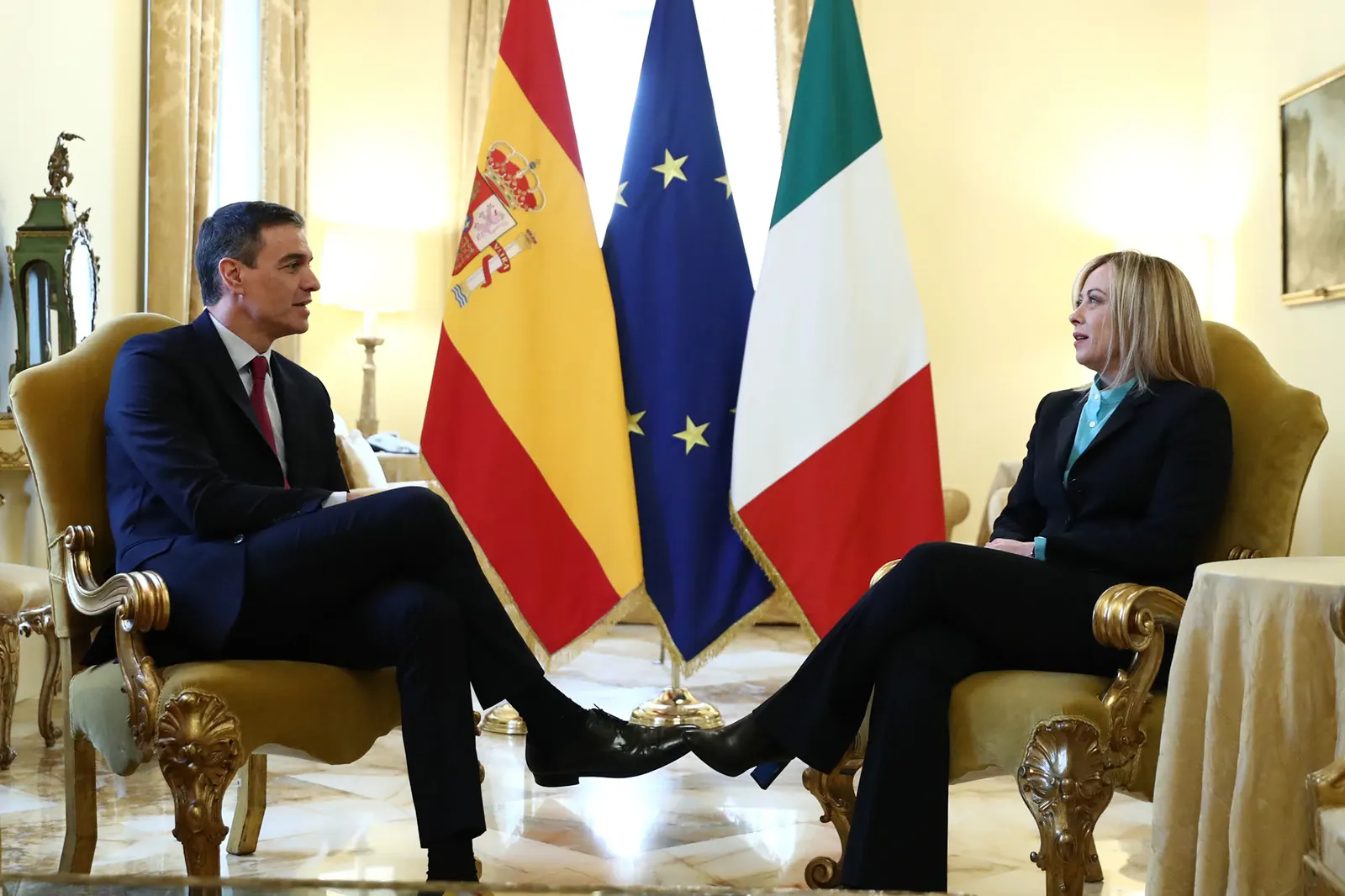 Pedro Sánchez y Giorgia Meloni, sentados frente a frente en sendos sillones dorados de un salón institucional, se miran mientras conversan; entre ambos, las banderas de España, la Unión Europea e Italia.