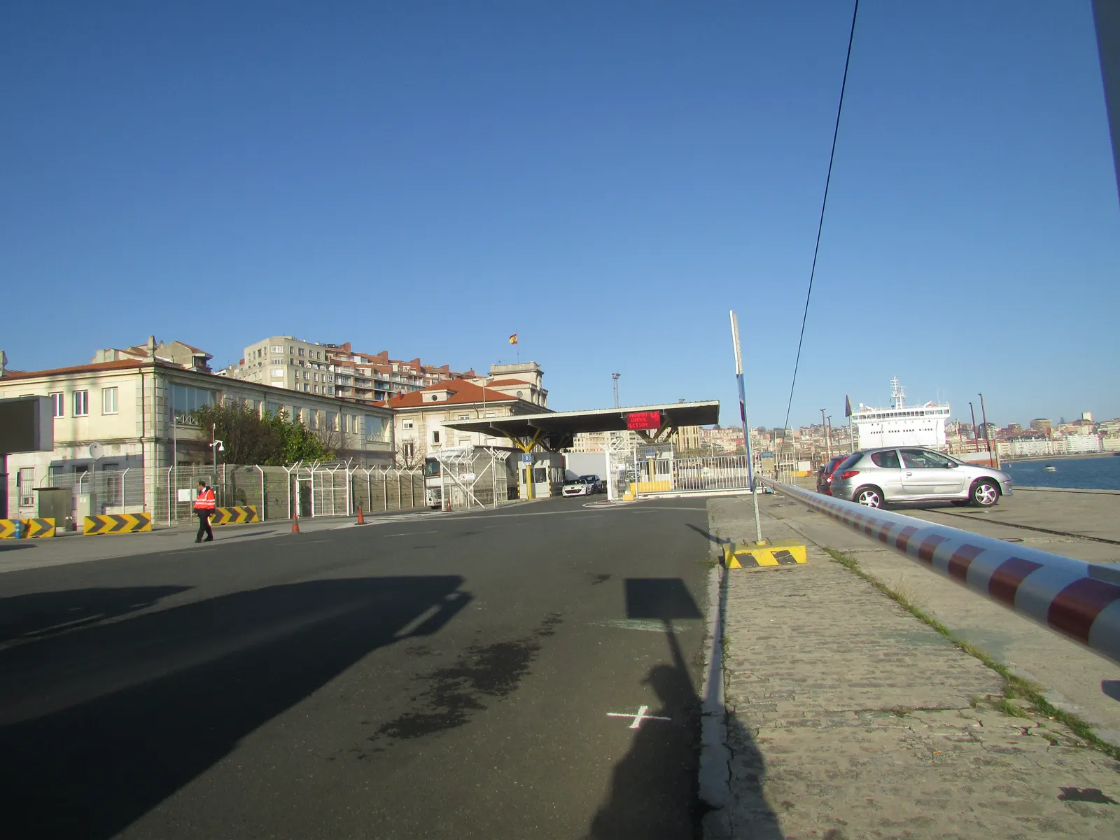 Puesto de control de pasaportes a la entrada de la terminal de ferris del puerto de Santander: una marquesina con un cartel rojo de «Passport control», vallas metálicas, coches en fila y un trabajador con chaleco reflectante; al fondo, un ferry atracado y la ciudad.