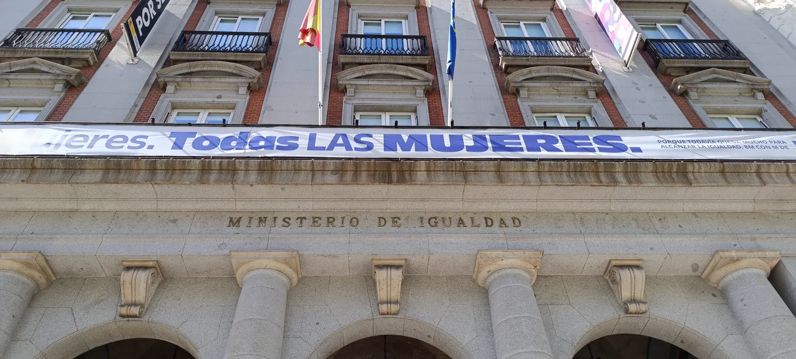 Fachada de la sede del Ministerio de Igualdad vista desde la acera: la inscripción «Ministerio de Igualdad» tallada en piedra sobre los arcos del soportal, las banderas de España y de la Unión Europea en los balcones y una pancarta institucional con el lema «Todas las mujeres».