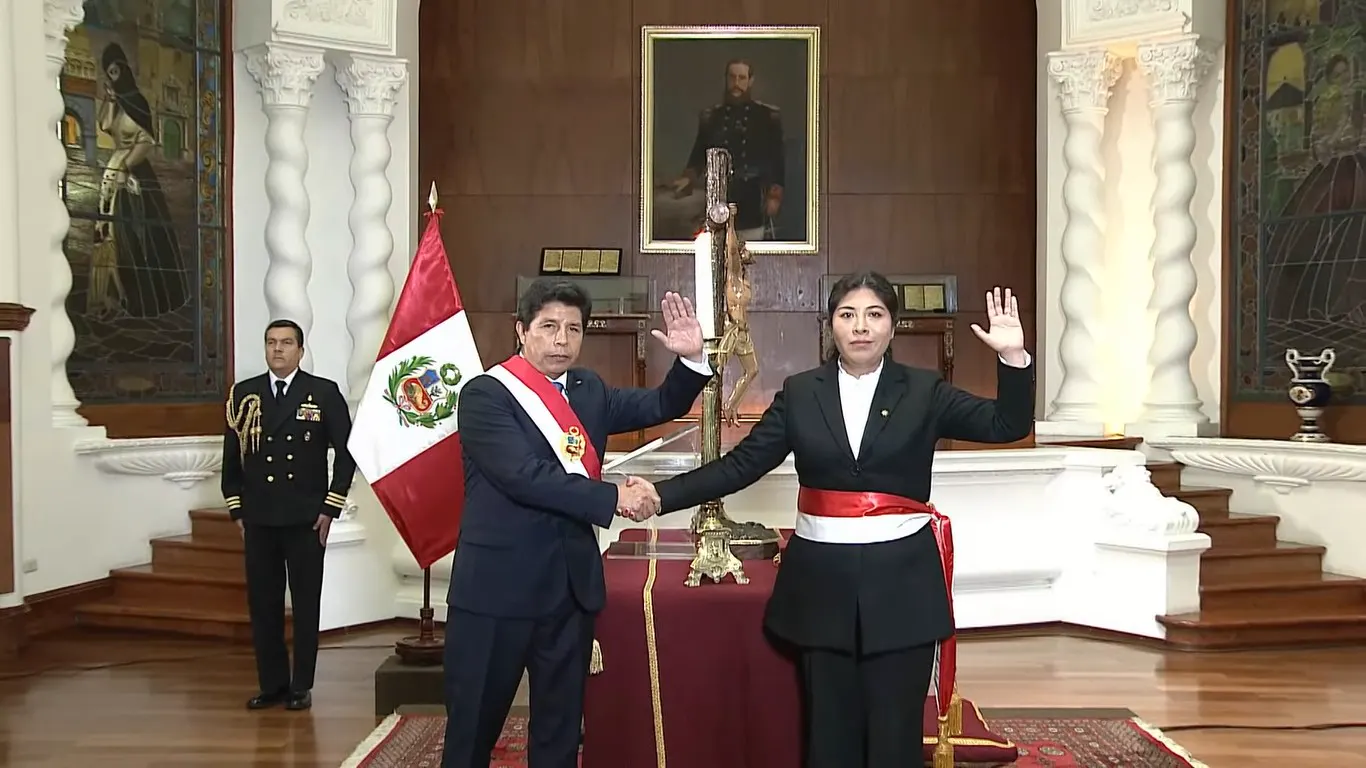Pedro Castillo, con la banda presidencial peruana, estrecha la mano de Betssy Chávez, que levanta la mano derecha para jurar el cargo, junto a la bandera de Perú en un salón del Palacio de Gobierno de Lima.