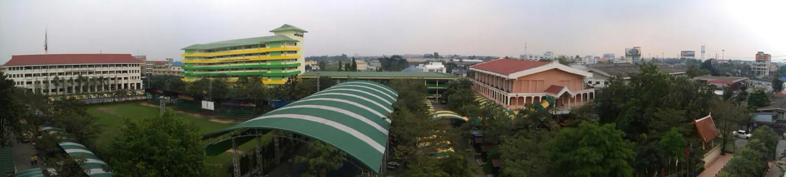 Vista panorámica del recinto del instituto Debsirin Nonthaburi: un edificio alargado de fachada verde y amarilla, otro de columnas rosadas, un pabellón cubierto con techo curvo verde y árboles alrededor, con la ciudad al fondo.