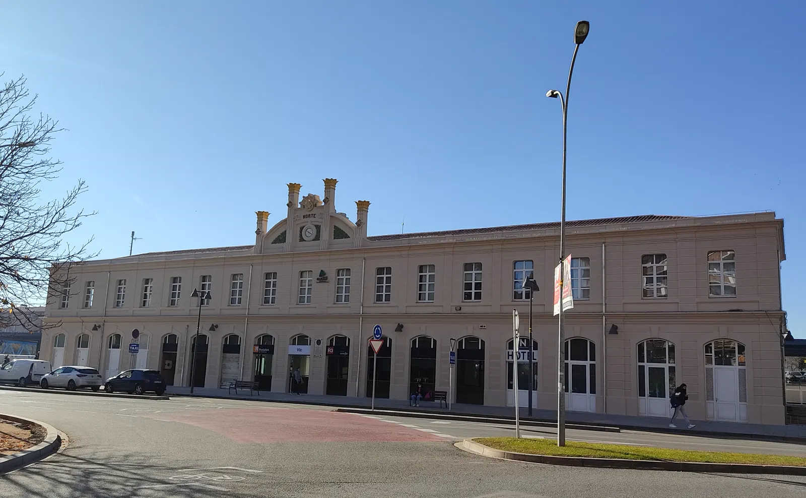 Fachada rehabilitada de la estación de tren de Vic, un edificio alargado de dos plantas con un frontón central con reloj, bajo un cielo azul despejado; junto a la puerta se leen los carteles de Rodalies y de la línea R3.