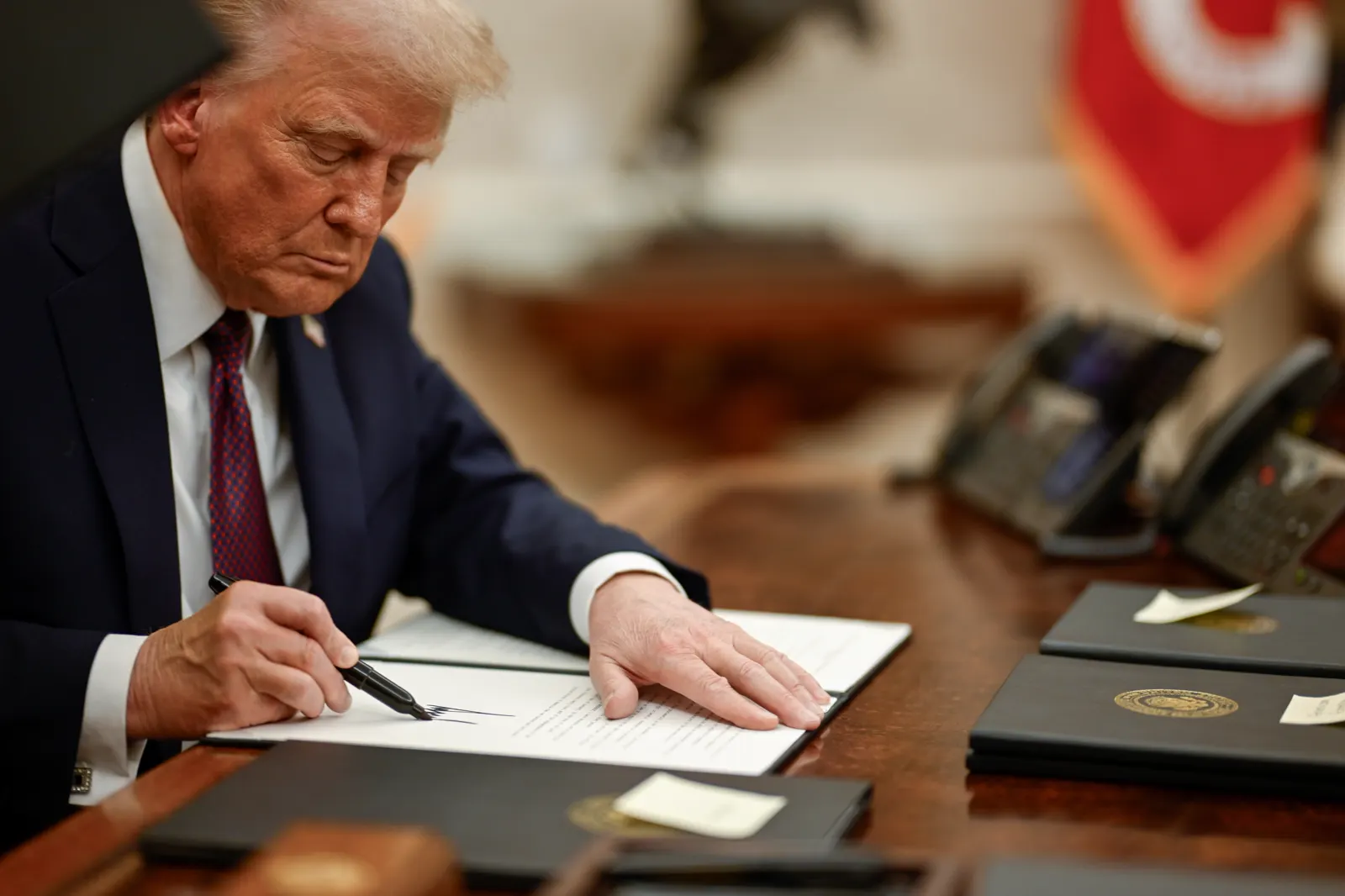 Donald Trump, sentado ante un escritorio de madera, firma con rotulador un documento oficial mientras sujeta el papel con la mano izquierda; a su lado, una pila de carpetas negras con el sello presidencial.
