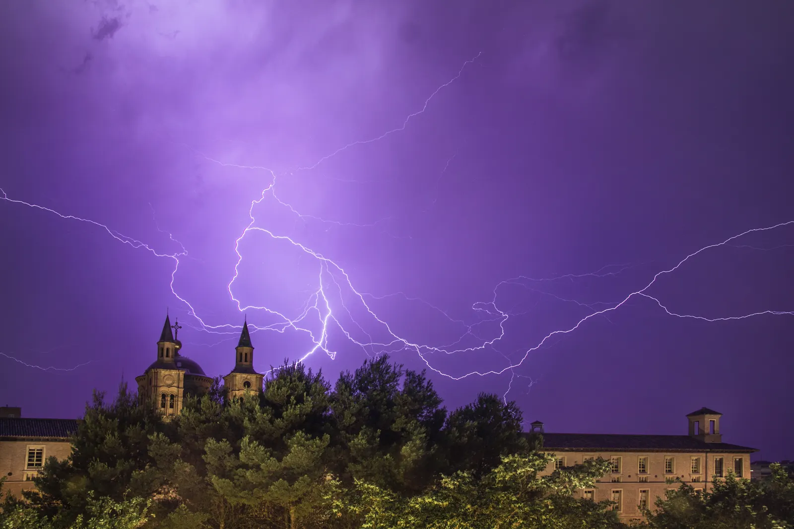 Varios rayos ramificados cruzan un cielo nocturno teñido de violeta sobre las torres de un edificio y una arboleda en Zaragoza.