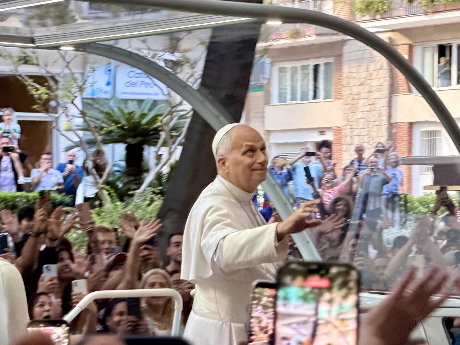 El papa León XIV, de blanco y con solideo, saluda con la mano derecha desde el papamóvil descubierto mientras avanza por una calle de Barcelona entre dos filas de personas que levantan los brazos y graban con sus teléfonos.
