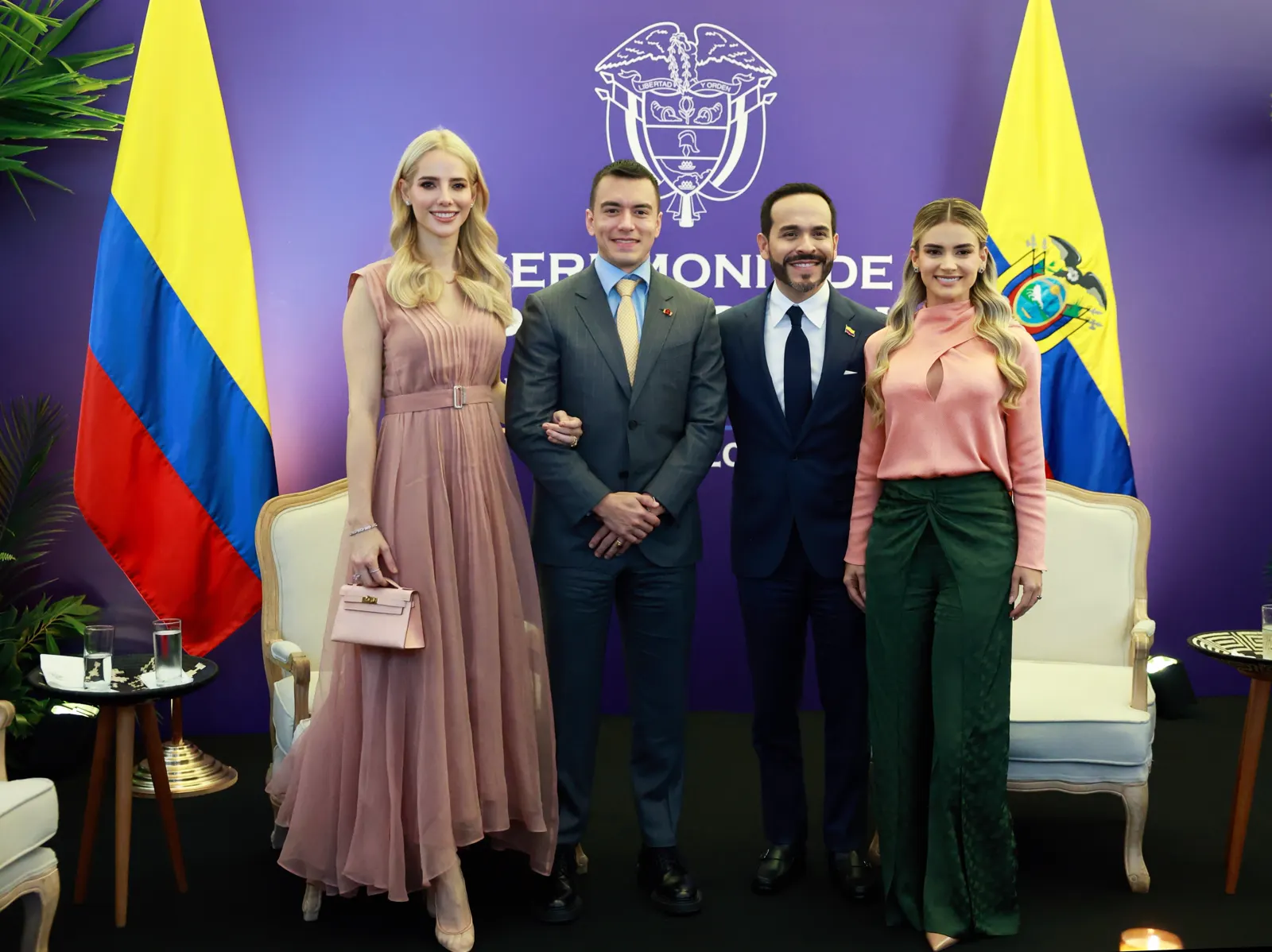 Cuatro personas posan sonrientes ante un panel morado con el escudo de Colombia, flanqueado por las banderas de Colombia y Ecuador; en el centro, dos hombres de traje oscuro cogidos del brazo.