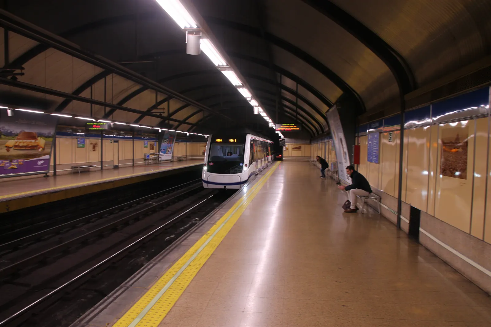 Andén subterráneo de una estación de metro con un tren blanco y azul entrando por la vía de la izquierda; dos viajeros esperan al fondo a la derecha, uno de ellos sentado en un banco.