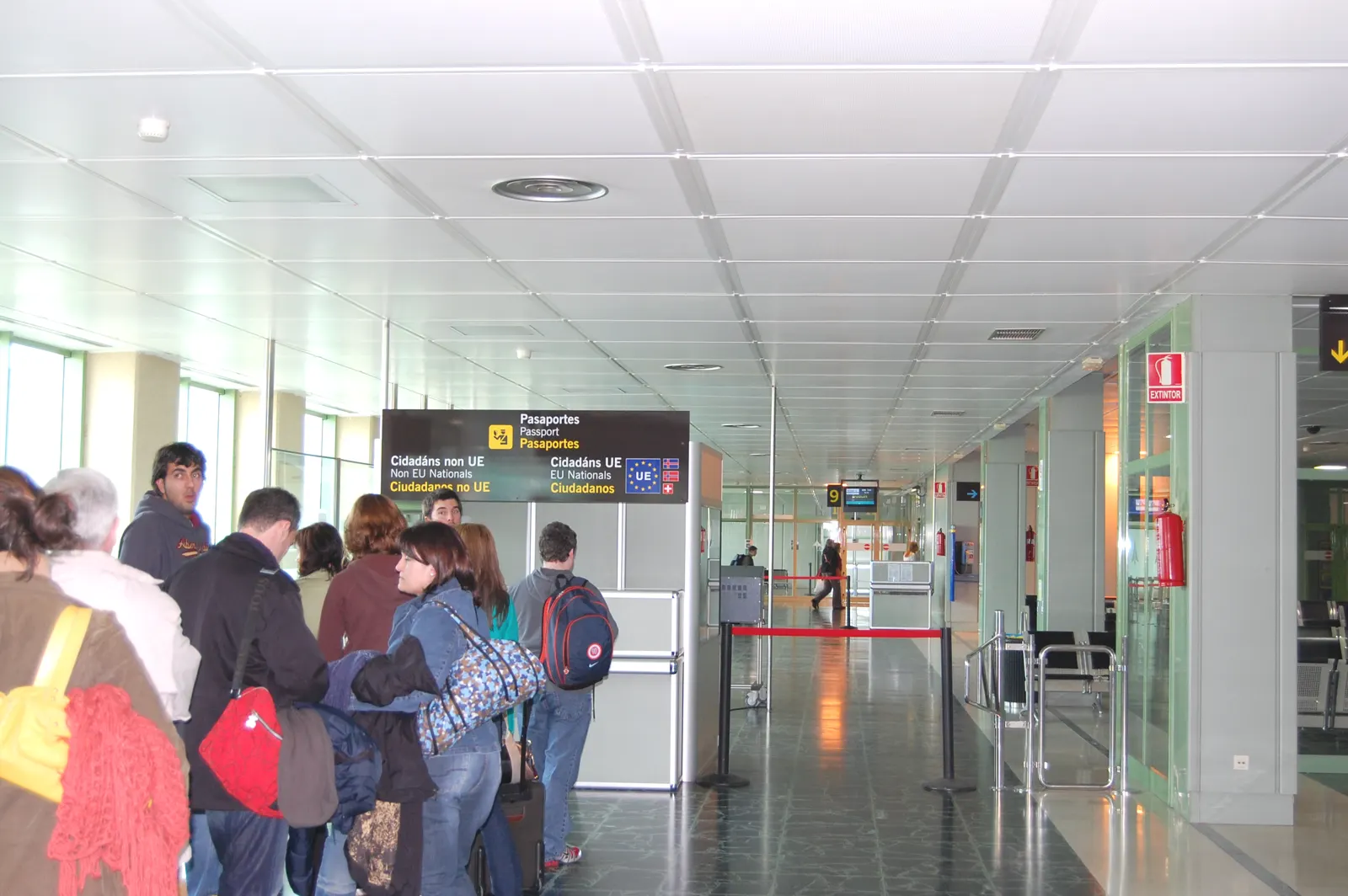 Cola de viajeros con equipaje de mano ante el control de pasaportes de un aeropuerto español, junto a un panel negro que separa dos carriles: «Cidadáns non UE / Non EU Nationals / Ciudadanos no UE» y «Cidadáns UE / EU Nationals / Ciudadanos UE».