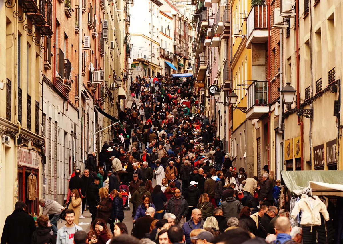 Calle estrecha del centro de Madrid vista desde arriba, abarrotada de personas que caminan entre puestos de venta ambulante, con fachadas de ladrillo y balcones a ambos lados.