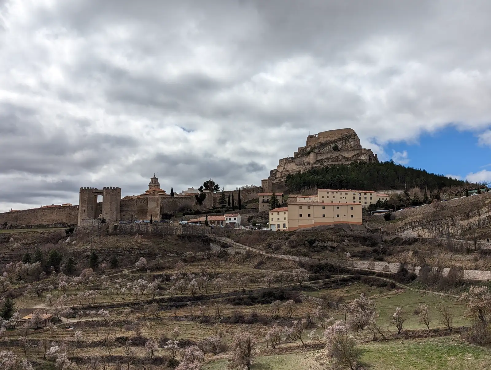 El pueblo amurallado de Morella y su castillo sobre un cerro rocoso, bajo un cielo cubierto de nubes grises y oscuras; en primer plano, bancales con almendros en flor.