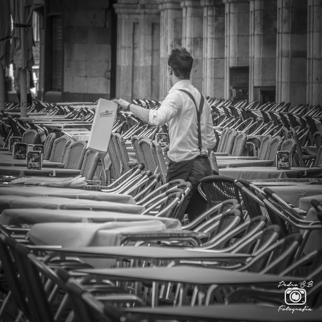 Fotografía en blanco y negro de un camarero de espaldas, con camisa blanca y tirantes, ordenando decenas de sillas y mesas apiladas en la terraza de un café bajo unos soportales