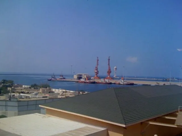 Vista del puerto de Jizán desde un tejado: grúas portuarias rojas y varios barcos amarrados en un muelle, con el mar Rojo y el cielo despejado al fondo.