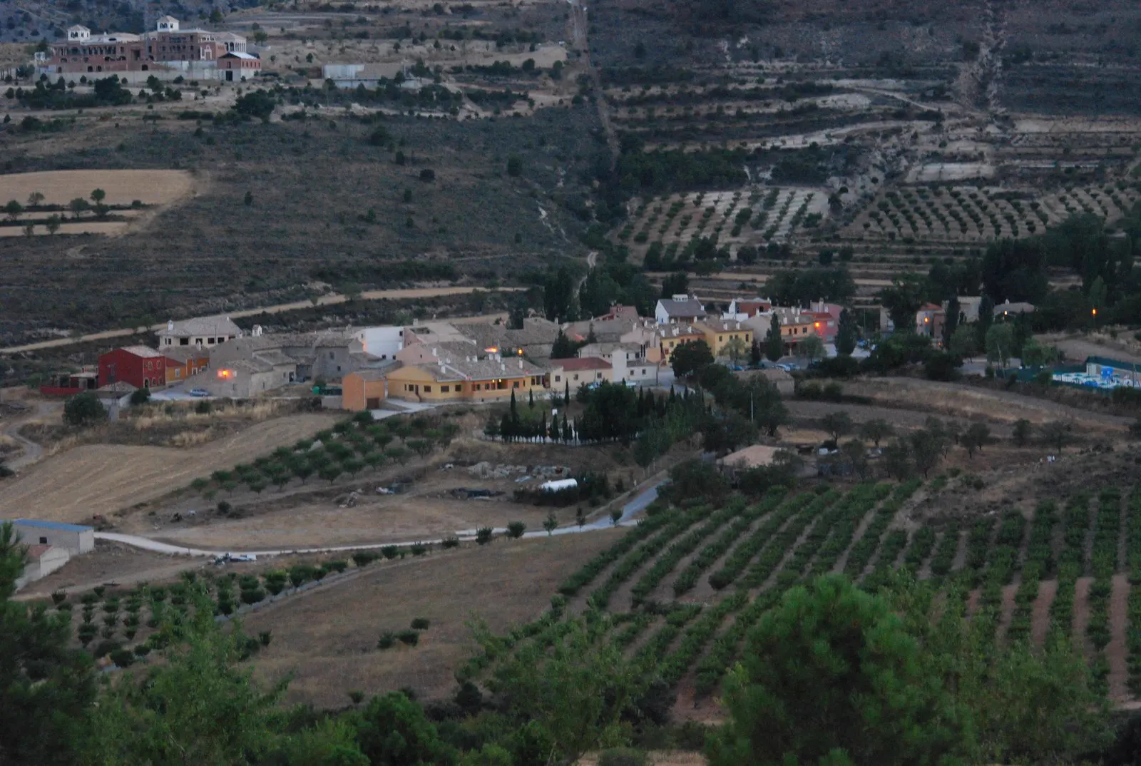 Vista aérea al atardecer de la pedanía de Inazares, un pequeño núcleo de casas de tejado rojizo en el fondo de un valle rodeado de bancales de almendros, viñedos, campos de cereal segados y laderas con pinar.