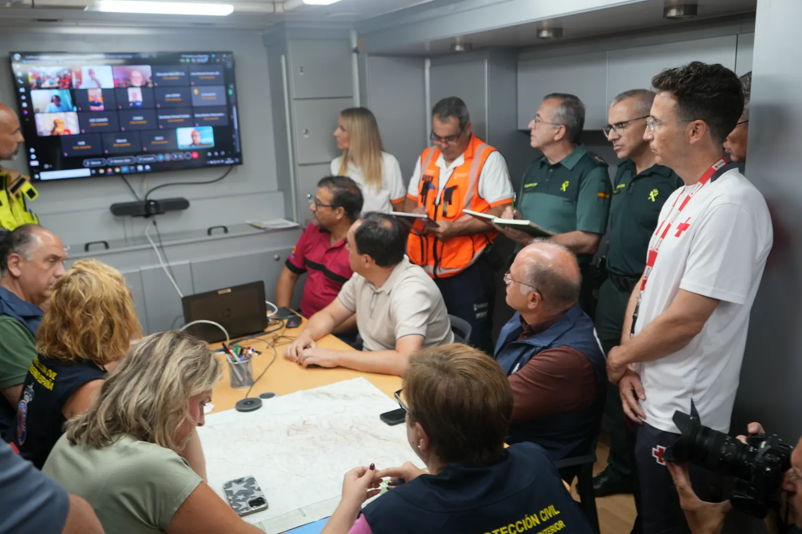 Reunión del Cecopi dentro de un vehículo de mando: una veintena de personas —guardias civiles de uniforme verde, técnicos con chaleco naranja, personal de Protección Civil y de Cruz Roja— rodean una mesa con un mapa topográfico desplegado, frente a una pantalla con una videoconferencia.