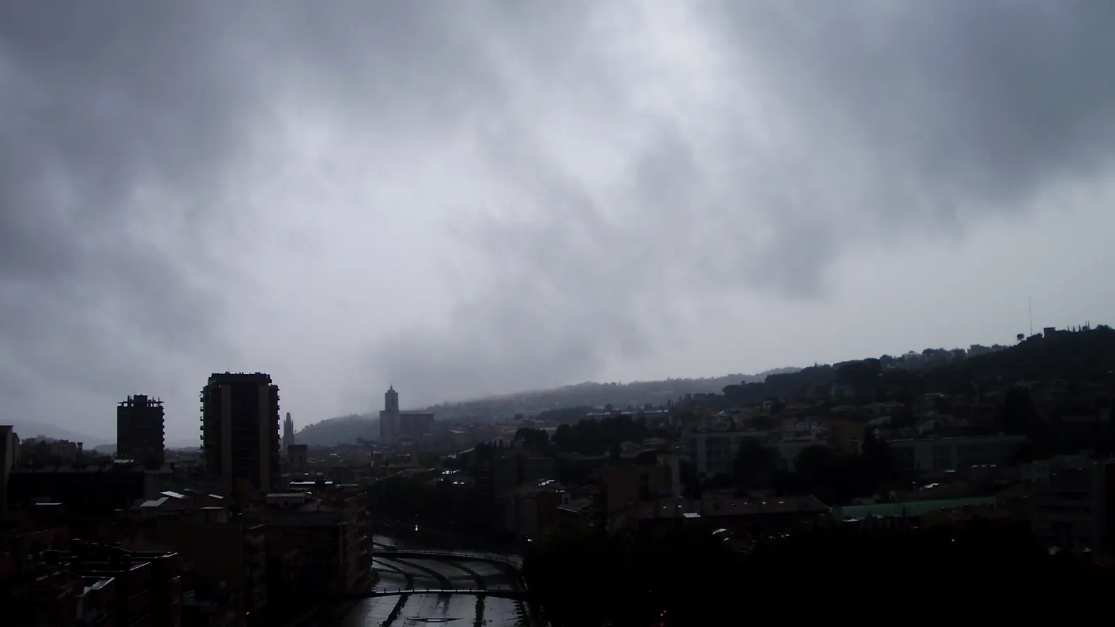 Nubes bajas y oscuras de tormenta cubriendo la ciudad de Girona; en el centro asoma la torre de la catedral y abajo discurre el río Onyar con sus puentes y las casas de colores.