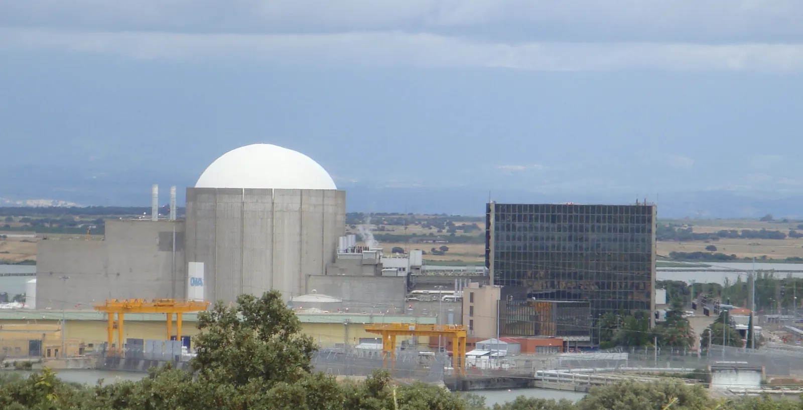 Vista de la central nuclear de Almaraz: la cúpula blanca del edificio de contención, los edificios de hormigón anexos y grúas amarillas, con el campo extremeño al fondo.