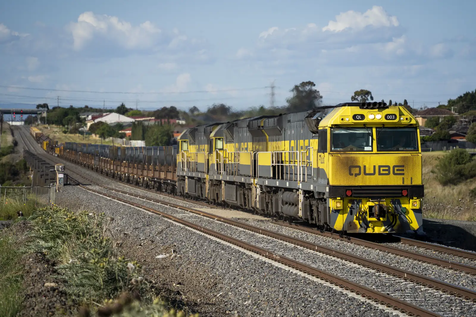 Tres locomotoras amarillas y grises con el rótulo QUBE arrastran una larga fila de vagones cargados de acero por una vía férrea, con casas y árboles al fondo