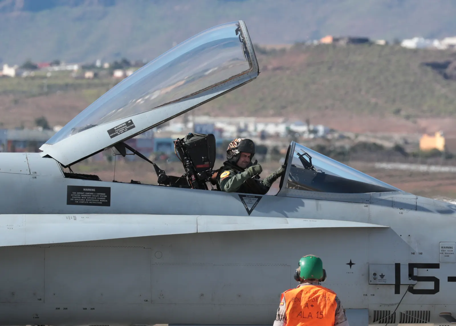 Piloto con casco y traje de vuelo, sentado en la cabina abierta de un caza F-18 del Ala 15 del Ejército del Aire español, hace una señal con la mano; en primer plano, de espaldas, un mecánico con casco verde y chaleco naranja con la inscripción «Ala 15» junto al fuselaje del avión.