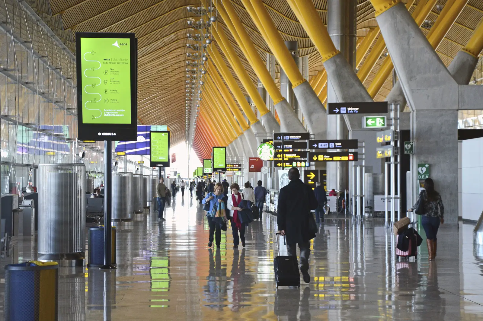 Pasillo interior de la terminal T4 del aeropuerto Adolfo Suárez Madrid-Barajas, con su techo ondulado de bambú y columnas amarillas; varios viajeros caminan con maletas bajo los paneles de señalización.