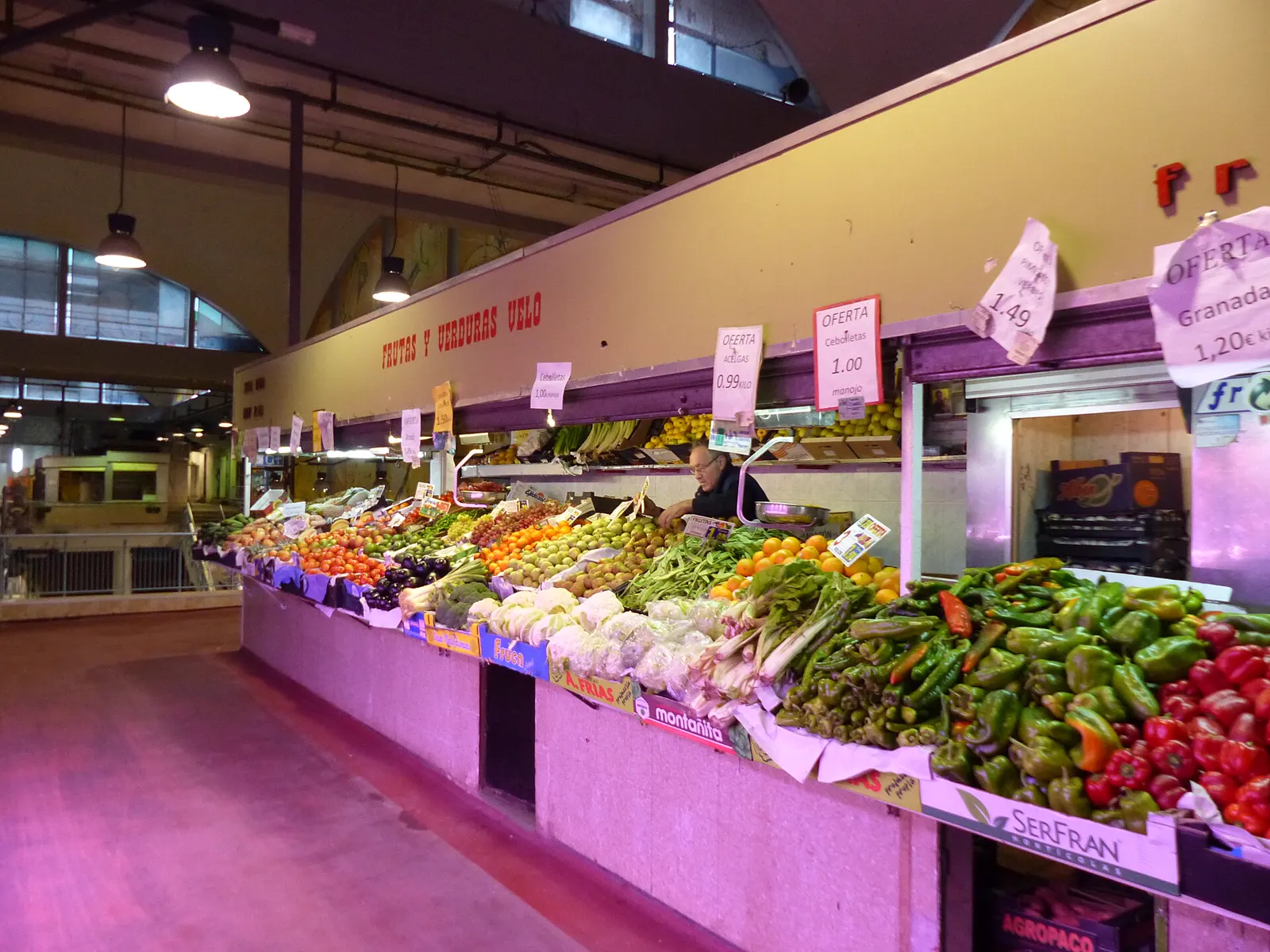 Puesto de frutas y verduras en un mercado cubierto, con cajones de pimientos, naranjas, tomates y hortalizas y carteles de oferta escritos a mano con los precios