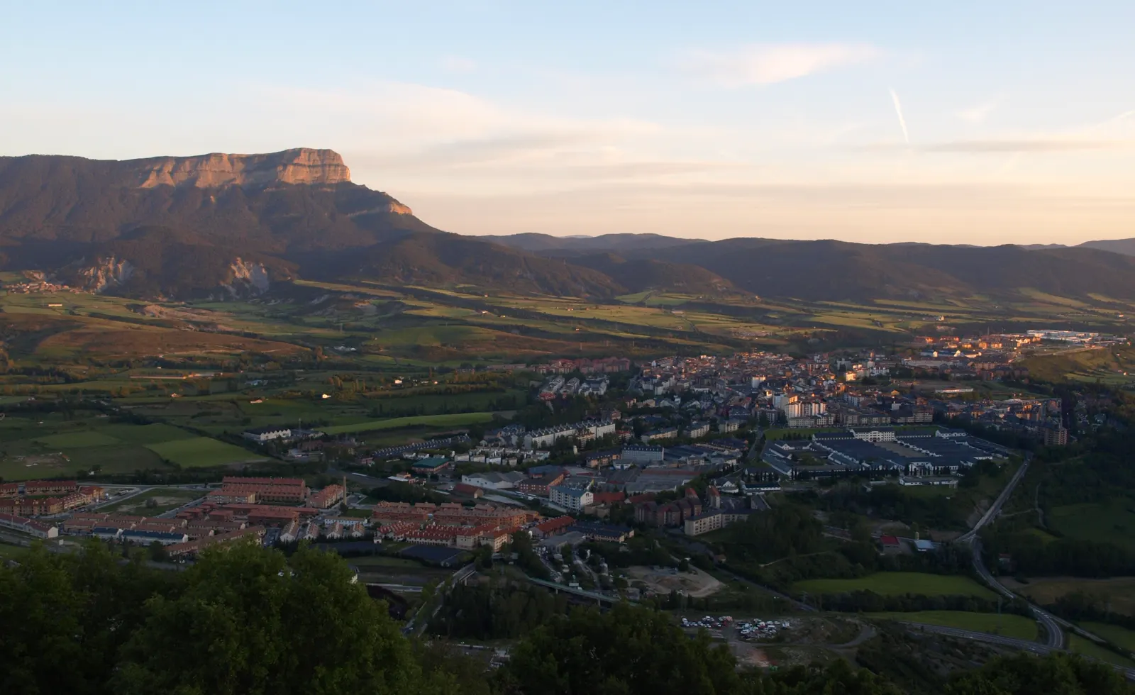 Vista de la ciudad de Jaca desde lo alto, con luz cálida de sol bajo: sus casas y los campos verdes del valle y, al fondo a la izquierda, la mole alargada y de cumbre plana de la Peña Oroel recortada contra el cielo.