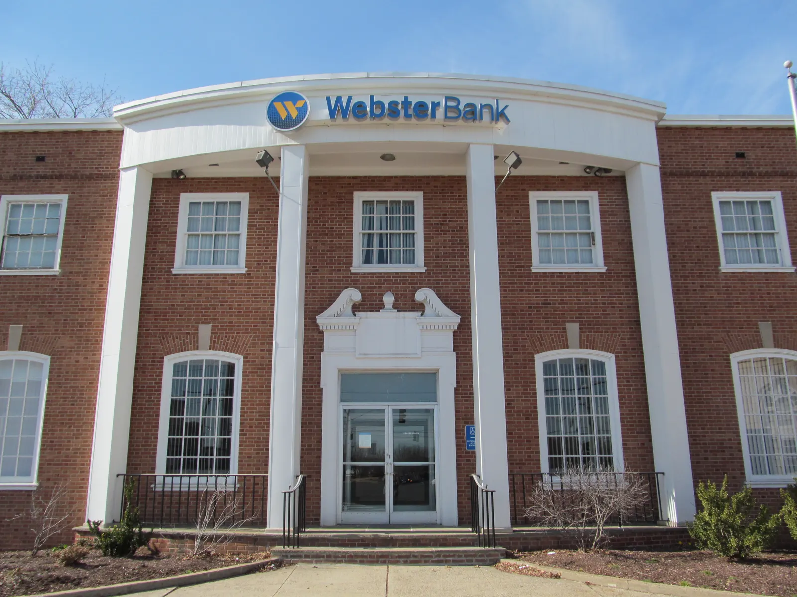 Fachada de ladrillo rojo de una oficina de Webster Bank, con pórtico blanco de columnas y el rótulo azul y amarillo del banco sobre la entrada, bajo un cielo despejado.