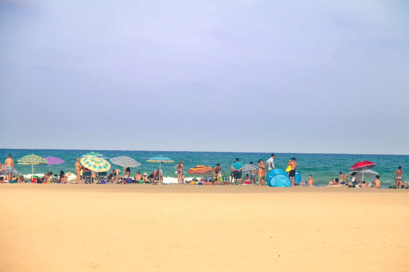Una franja ancha de arena vacía en primer plano y, al fondo, junto a la orilla del mar, un grupo de bañistas con sombrillas de colores y una tienda de playa azul bajo un cielo despejado.