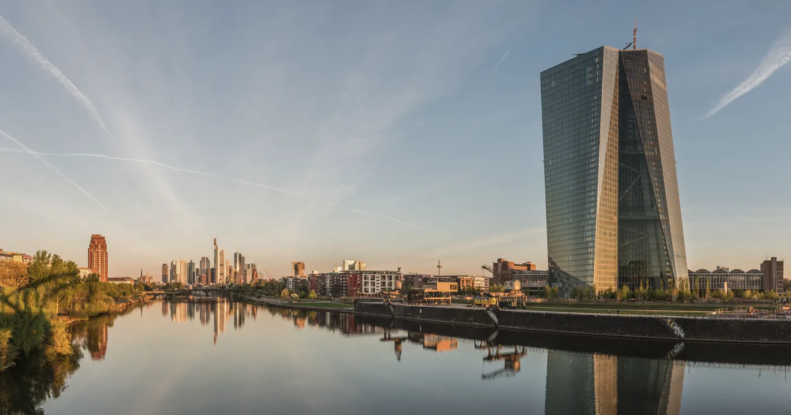 La torre de cristal del Banco Central Europeo, junto al río Meno, reflejada en el agua al amanecer, con el perfil de rascacielos de Fráncfort al fondo