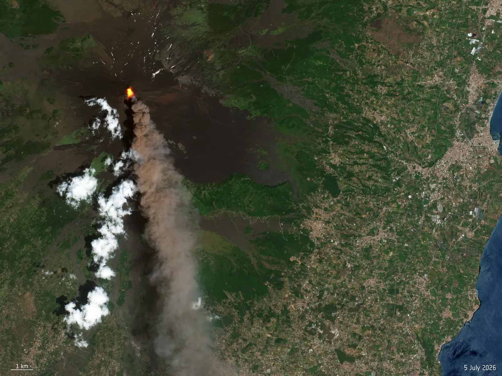 Imagen de satélite del Etna en erupción: un punto de lava incandescente en la cumbre y una larga columna de ceniza gris que se extiende hacia el sureste sobre el flanco del volcán, con la llanura poblada y la costa de Sicilia a la derecha.
